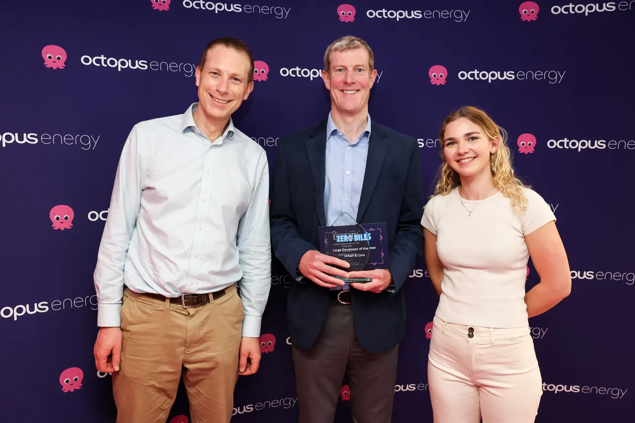 Three people smiling and standing in front of a purple Octopus Energy branded backdrop. The man in the center wears a dark blue blazer and holds a clear glass trophy with text confirming Urban&Civic has won an Octopus Energy Zero Bills Award as "Large Developer of the Year